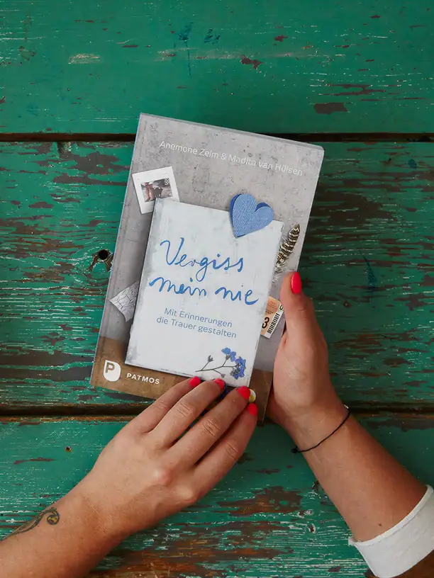 Zwei Hände halten das Buch Vergiss mein nie von Anemone Zeim und Madita van Hülsen über einem grün gestrichenen Holztisch